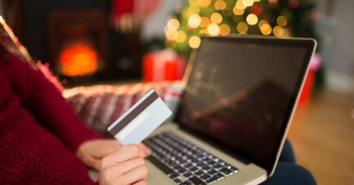 A closeup of a woman's hand holding a debit card in front of an open laptop computer, with a Christmas tree and presents in the background